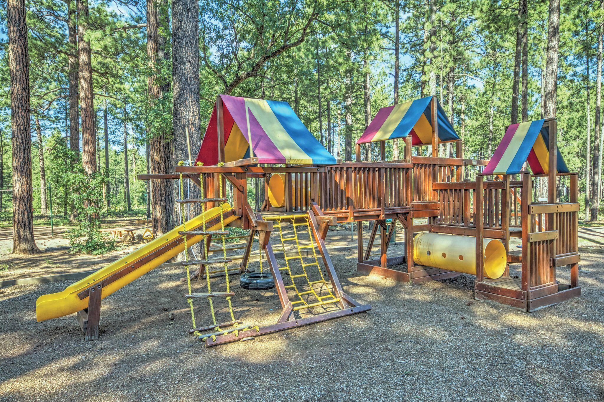 A playground with a slide, climbing net, and a tire swing in a forested area.