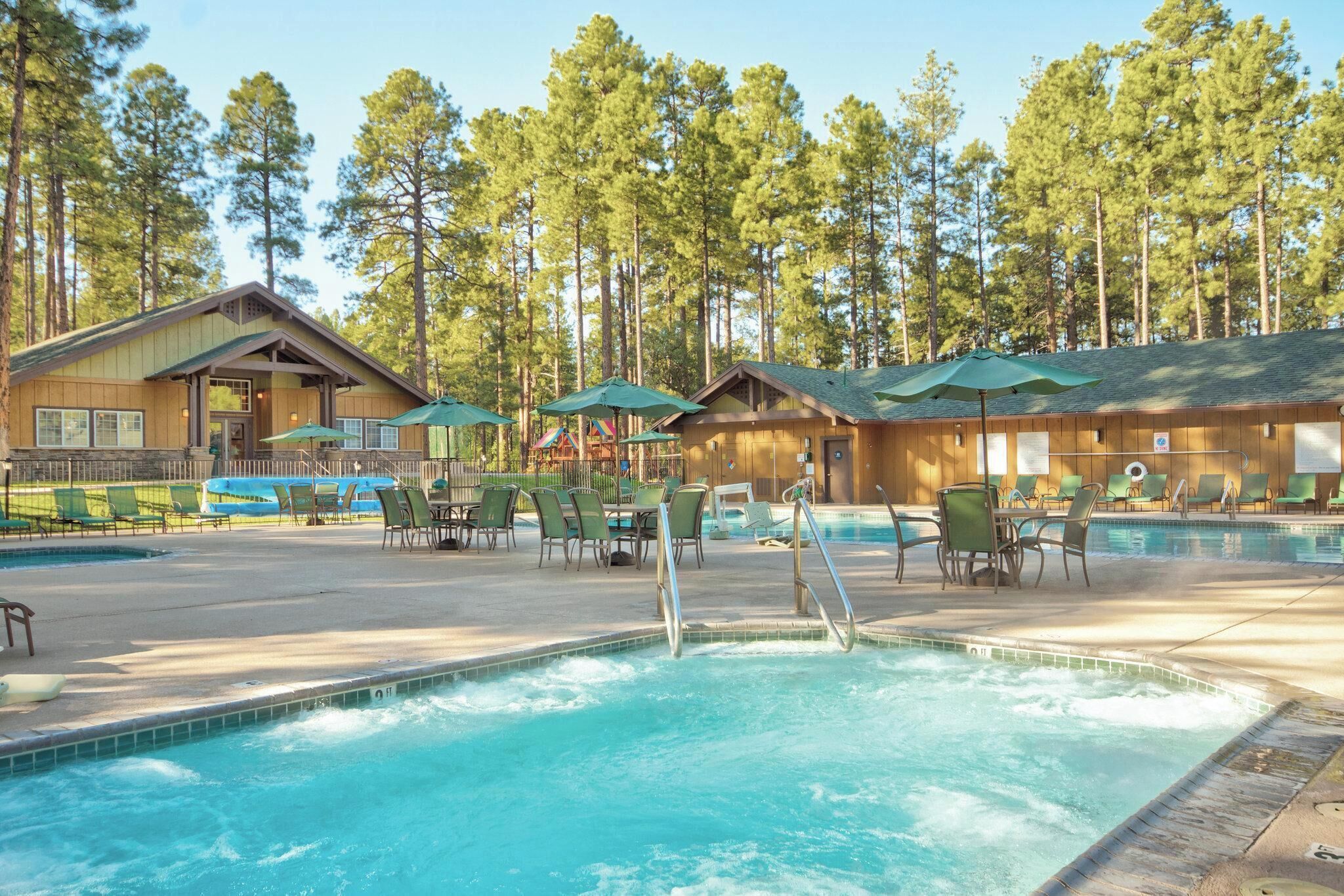A swimming pool with lounge chairs and umbrellas, surrounded by wooden cabins and tall trees.