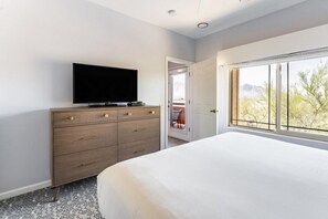 A bedroom with a bed, a dresser, a flat-screen TV, and a window with a view of trees and mountains. - Oro Valley | Group Sonoran Retreat | Pool + Hot Tub | 3BR Sleeps 6 (Oro Valley)