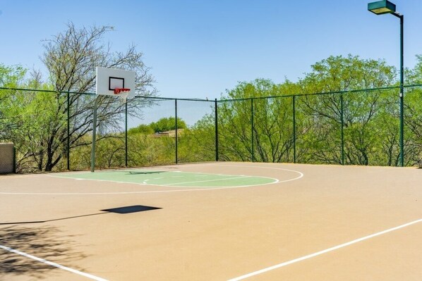 An outdoor basketball court with a hoop, surrounded by a chain-link fence and trees.