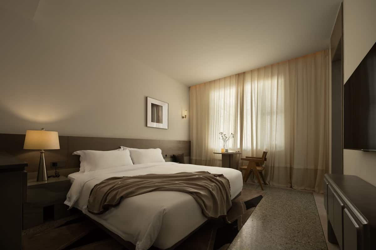 A hotel room with a bed, bedside table, lamp, chair, desk, and a TV on the wall.