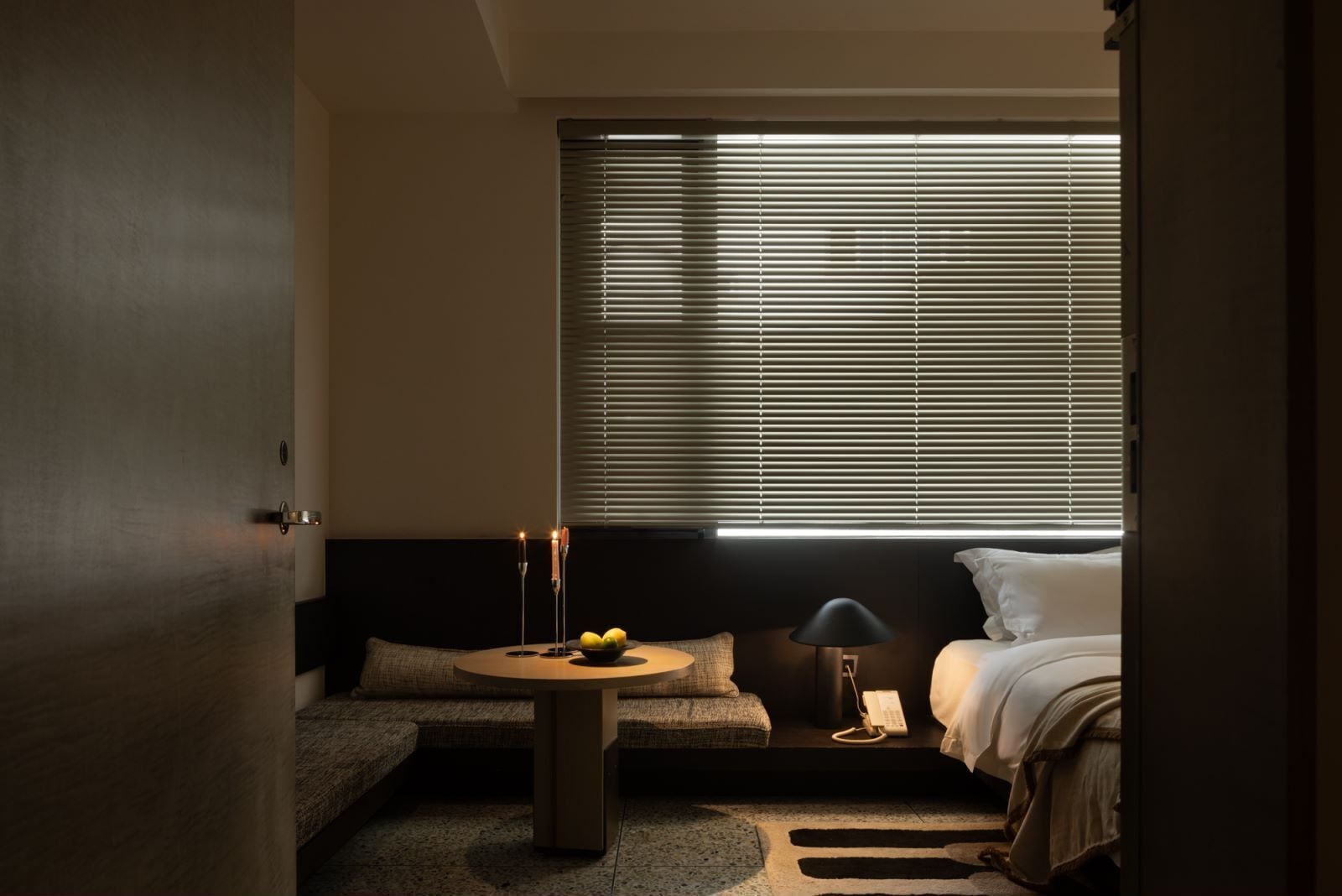 A hotel room with a bed, a small round table with a bowl of fruit, a lamp, and a telephone.