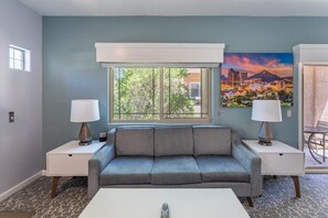 A living room with a gray sofa, two nightstands with lamps, a window with a view of trees and buildings, and a painting of a cityscape on the wall. - Oro Valley | Couples Desert Escape | Pool + Hot Tub (Oro Valley)
