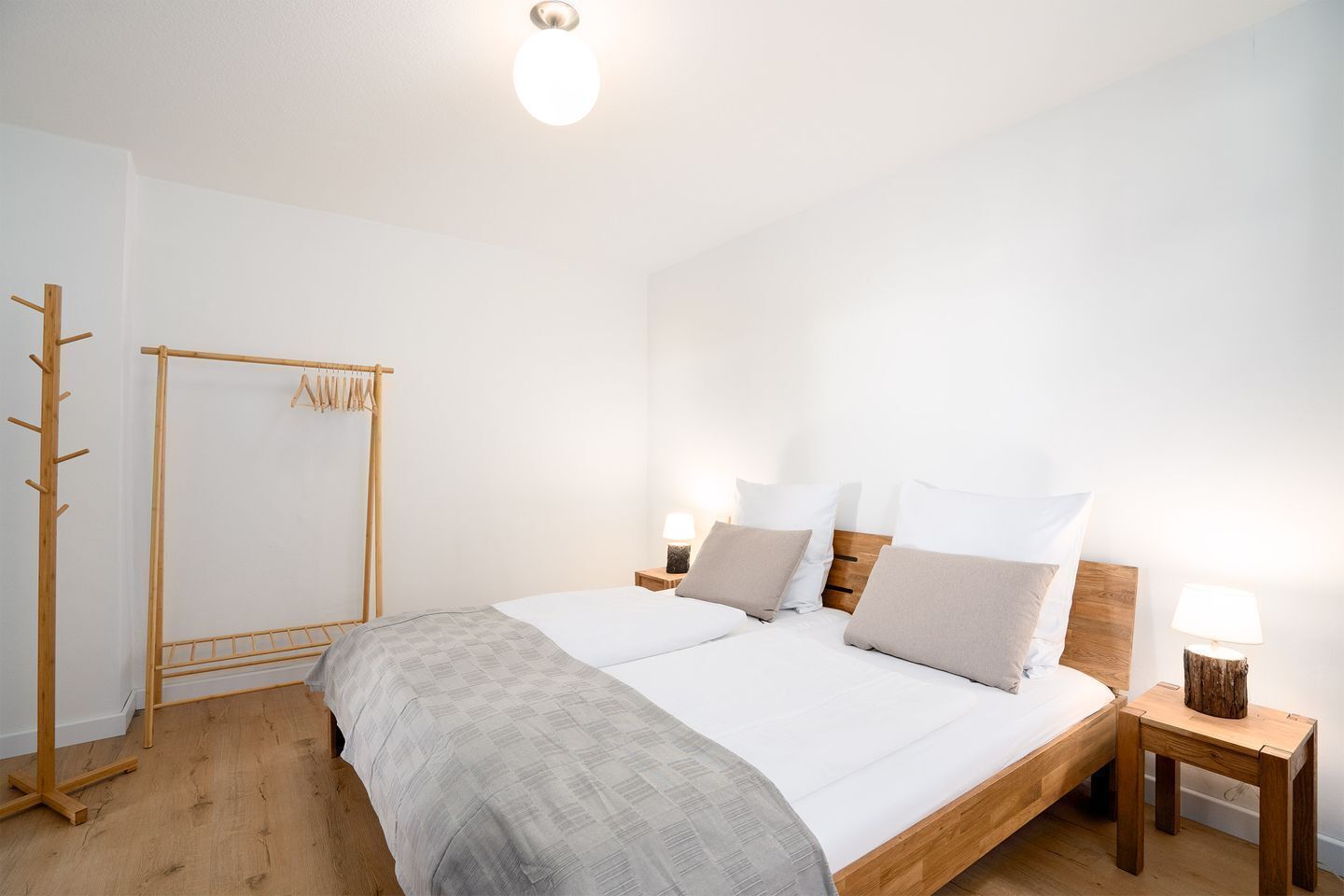 Un dormitorio con cama de madera, dos mesitas de noche con lámparas, suelo de madera y un tendedero.