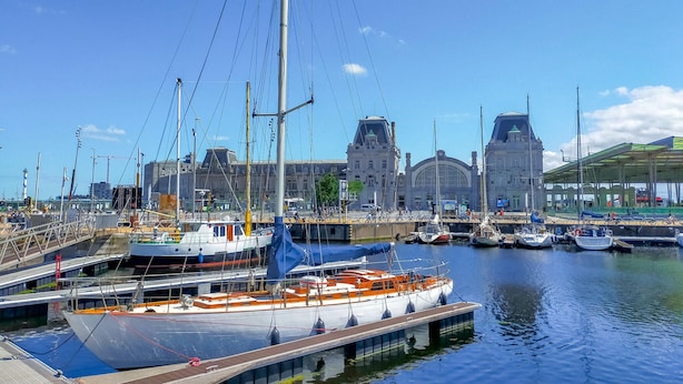 Een jachthaven met afgemeerde boten, een groot gebouw op de achtergrond en een voetgangersbrug.