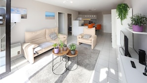 Een moderne woonkamer met een bank, twee fauteuils, een salontafel en een televisie. In de achtergrond is een keuken met een magnetron en een koelkast.