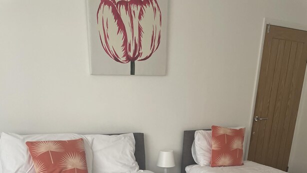 A hotel room with two beds, white bedding, and red patterned pillows. There is a grey towel on the bed and a wooden door in the background. - 3Bed House, Sleeps 6 near Luton Airport w/ Parking (Luton)