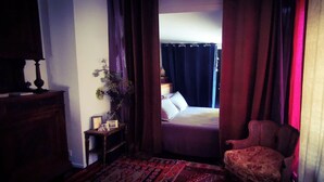 A room with a bed, a chair, a small table with a lamp, and a plant.