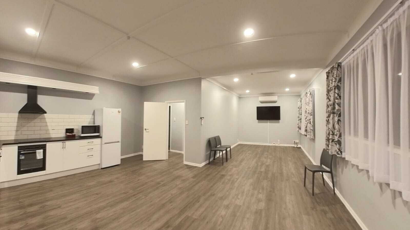 Un salón amplio con cocina, una mesa de comedor con sillas, un televisor de pantalla plana montado en la pared y un gran ventanal con cortinas.
