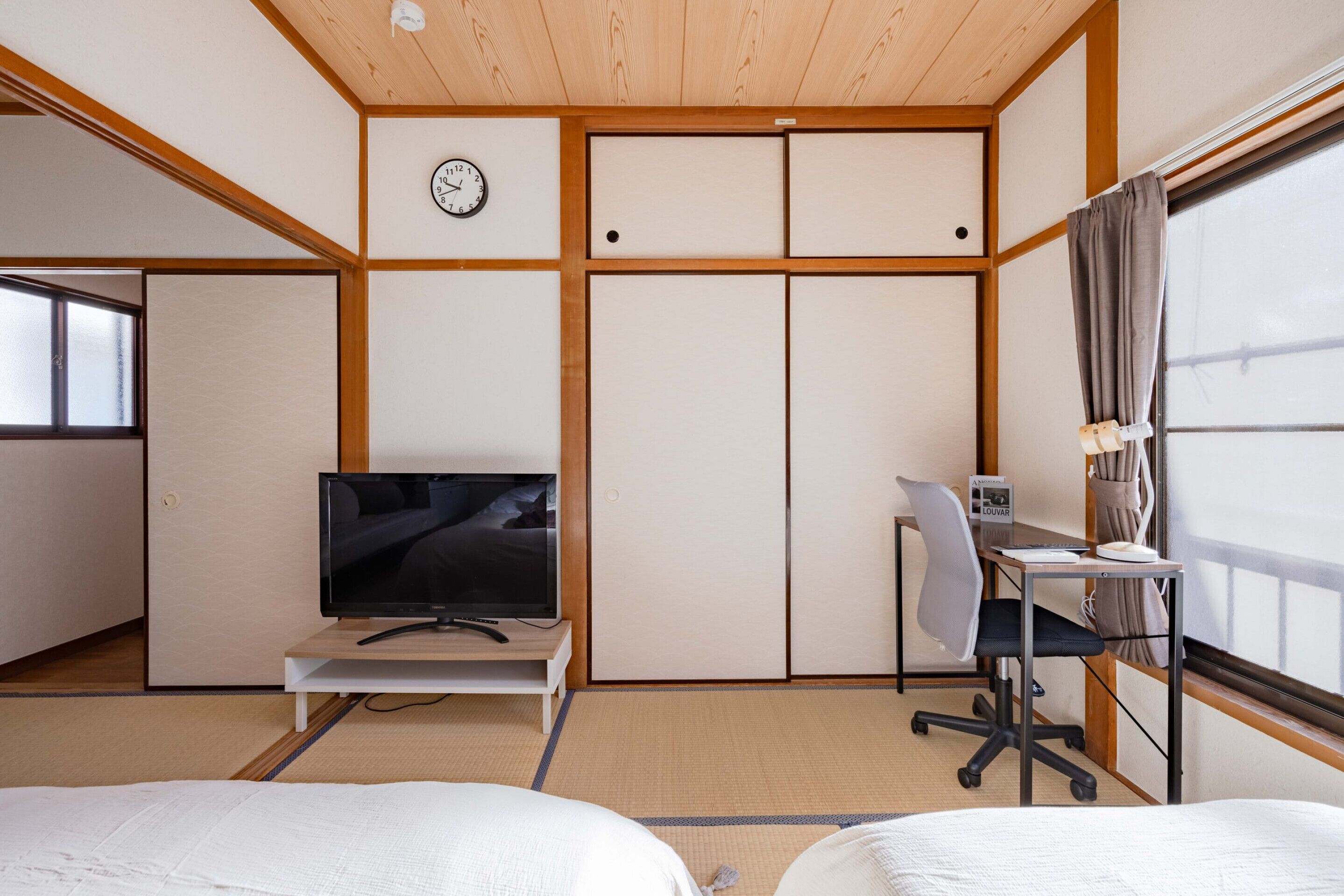 A room with tatami flooring, a television on a low table, a desk with a chair, and a large window with curtains.