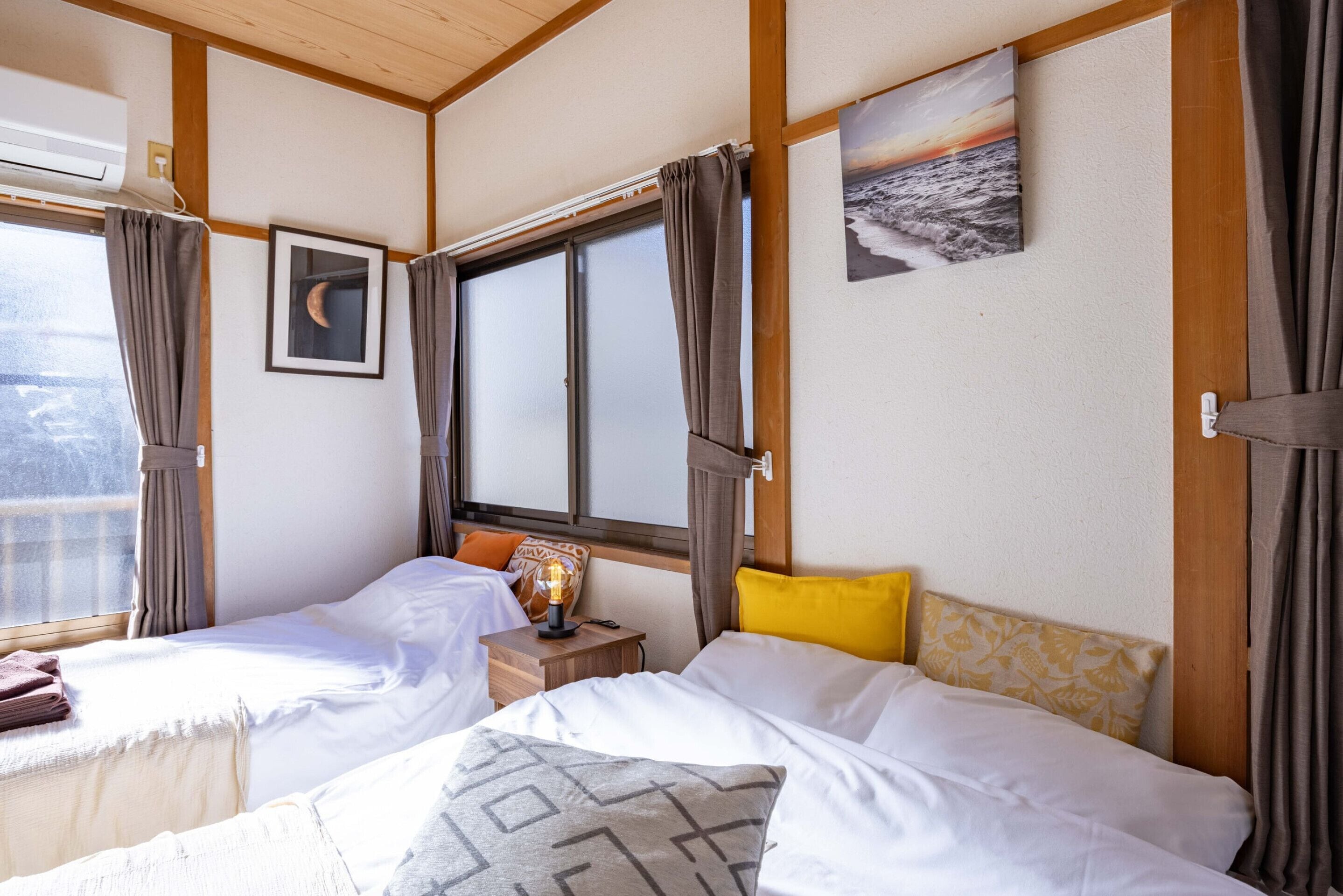 Een hotelkamer met twee bedden, een raam met gordijnen, een nachtkastje met lamp en een schilderij van een zonsondergang aan de muur.