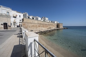 Un paseo marítimo con un muro de piedra, mar azul cristalino y edificios blancos.