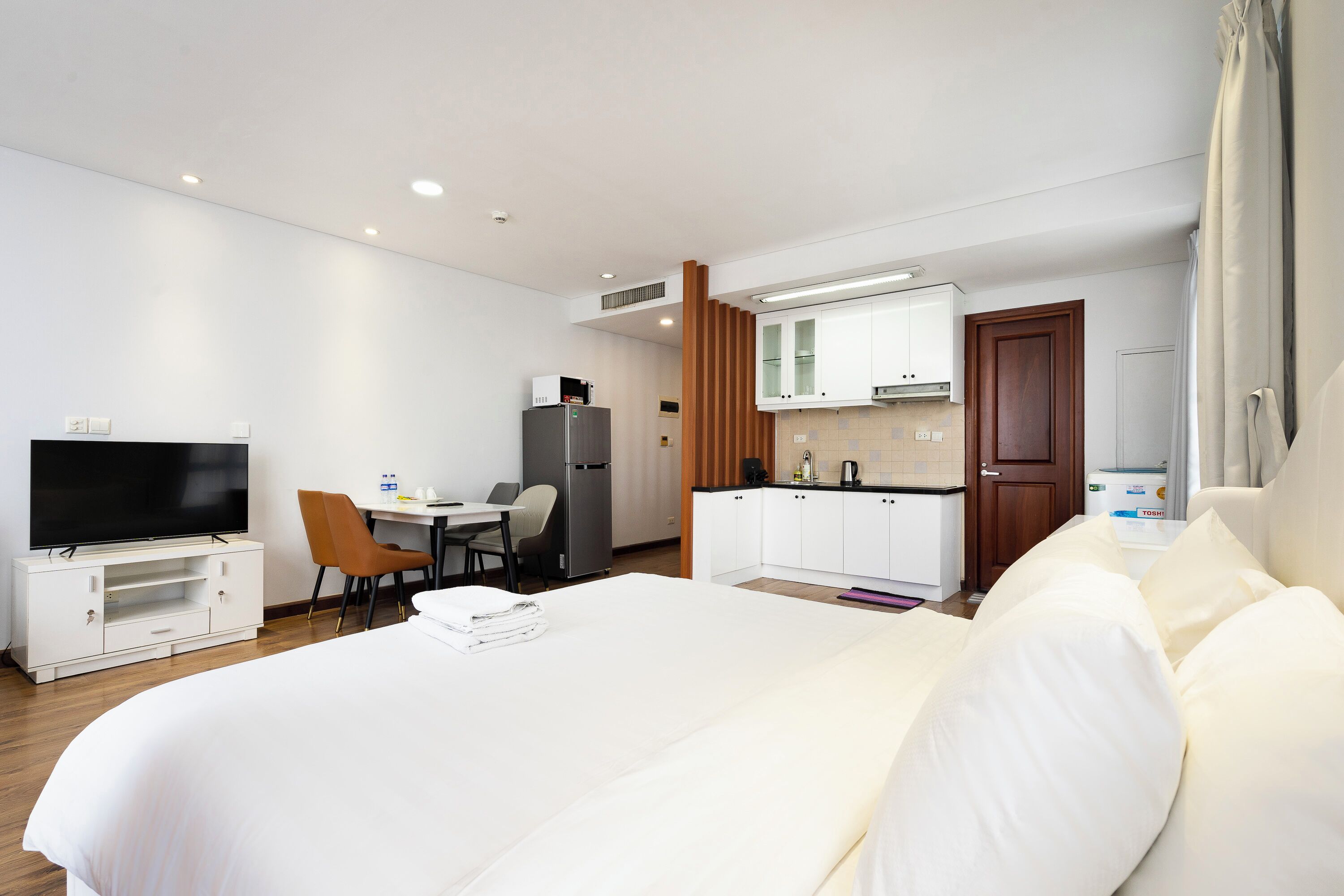 A hotel room with a large bed, a TV, a desk with a chair, a kitchenette, and a bathroom.