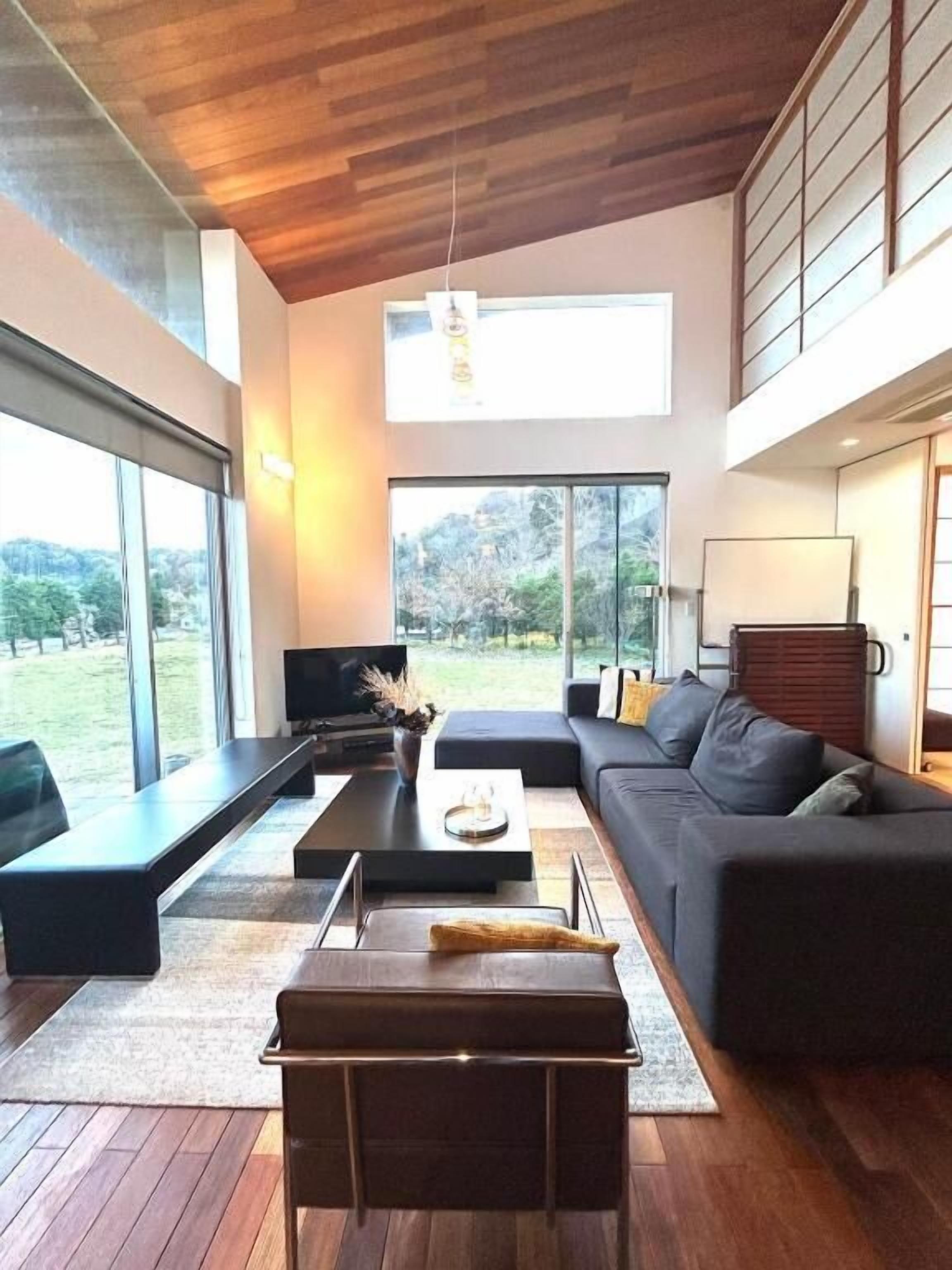 A modern living room with a sofa, coffee table, and ottoman, large windows, and a television.