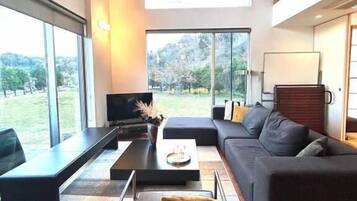 A modern living room with a sofa, coffee table, and ottoman, large windows, and a television.