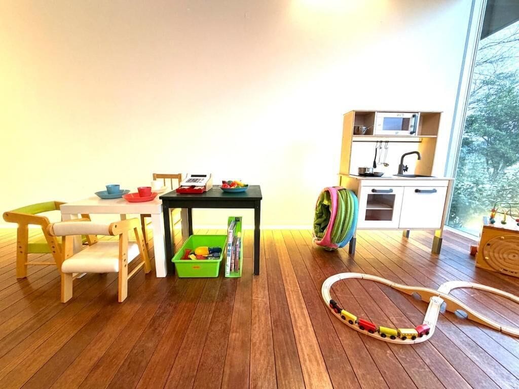 A room with a wooden floor, a table with chairs, a green storage bin, a toy train set, and a window with a view of trees.