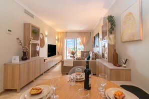 Un salon moderne avec un canapĂ©, une table Ă manger agrĂ©mentĂ©e de verres Ă vin et dâassiettes, un tĂ©lĂ©viseur et une vue sur lâextĂ©rieur.