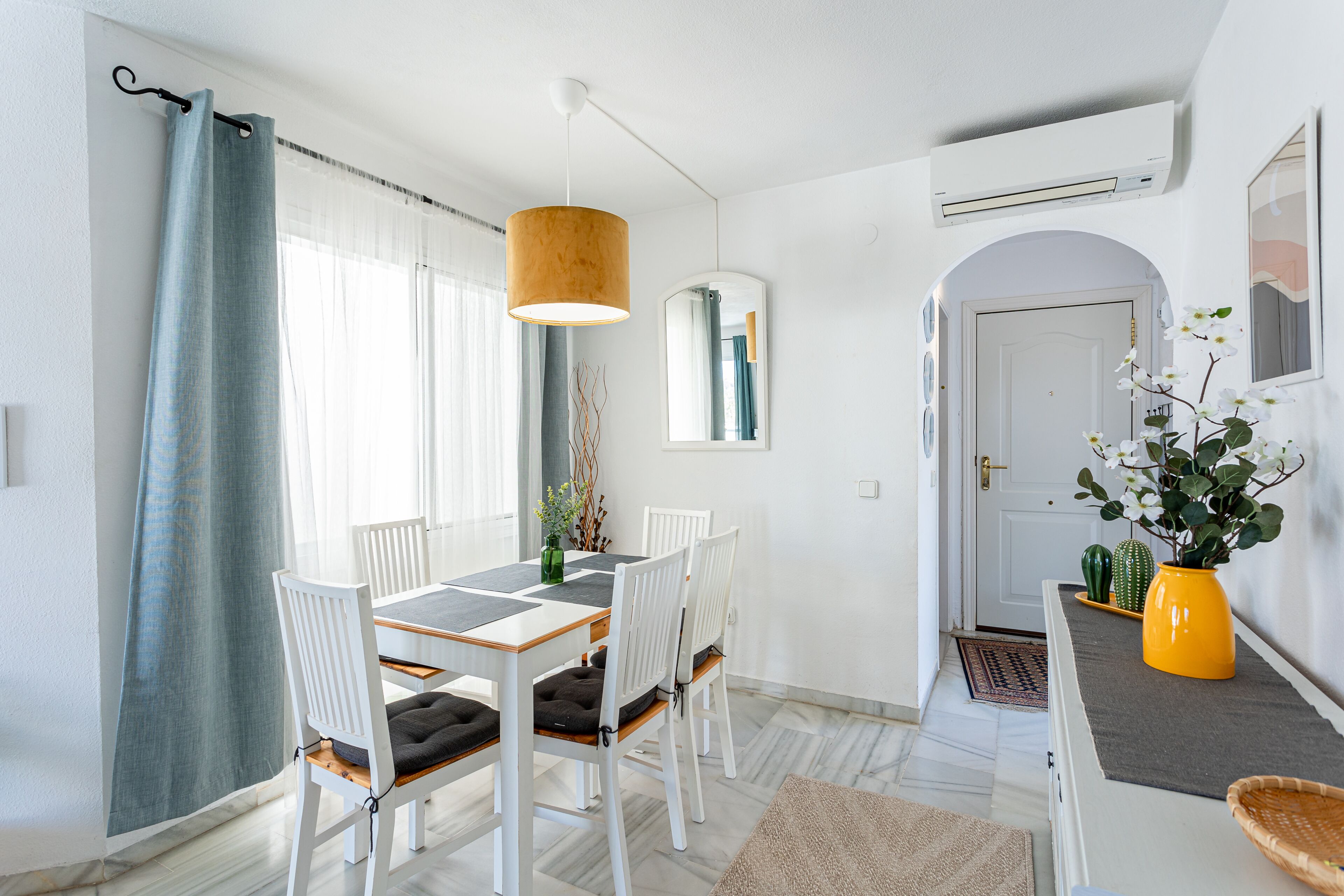 Un coin repas avec une table et des chaises blanches, un luminaire suspendu et un miroir au mur.