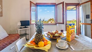 A room with a bed, a desk with a computer, a breakfast table with fruits, a carton of Cornflakes, and a view of the sea. - 1 Le Perle Di Stintino, Stintino, Italy (Stintino)