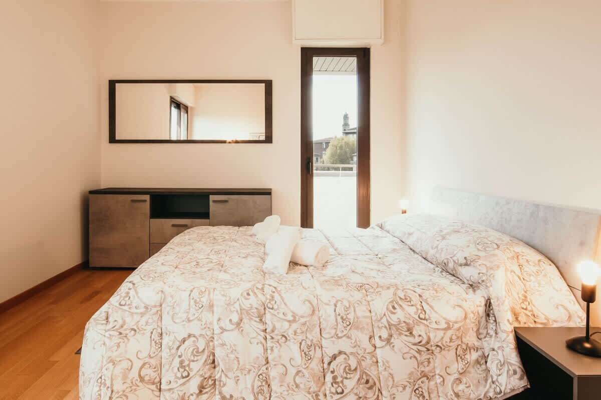 Un dormitorio con una cama, una mesita de noche con lámpara, un aparador y un espejo grande en la pared.