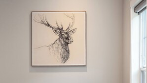 Ett modernt vardagsrum med ett stort, inramat konstverk föreställande ett rådjur på väggen, en mörk lädersoffa, ett litet sidobord med en växt och ett fönster som släpper in naturligt ljus.