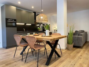 Une cuisine moderne avec des meubles de couleur sombre, un four encastré et un coin repas agrémenté de chaises en osier et d’une table en bois.