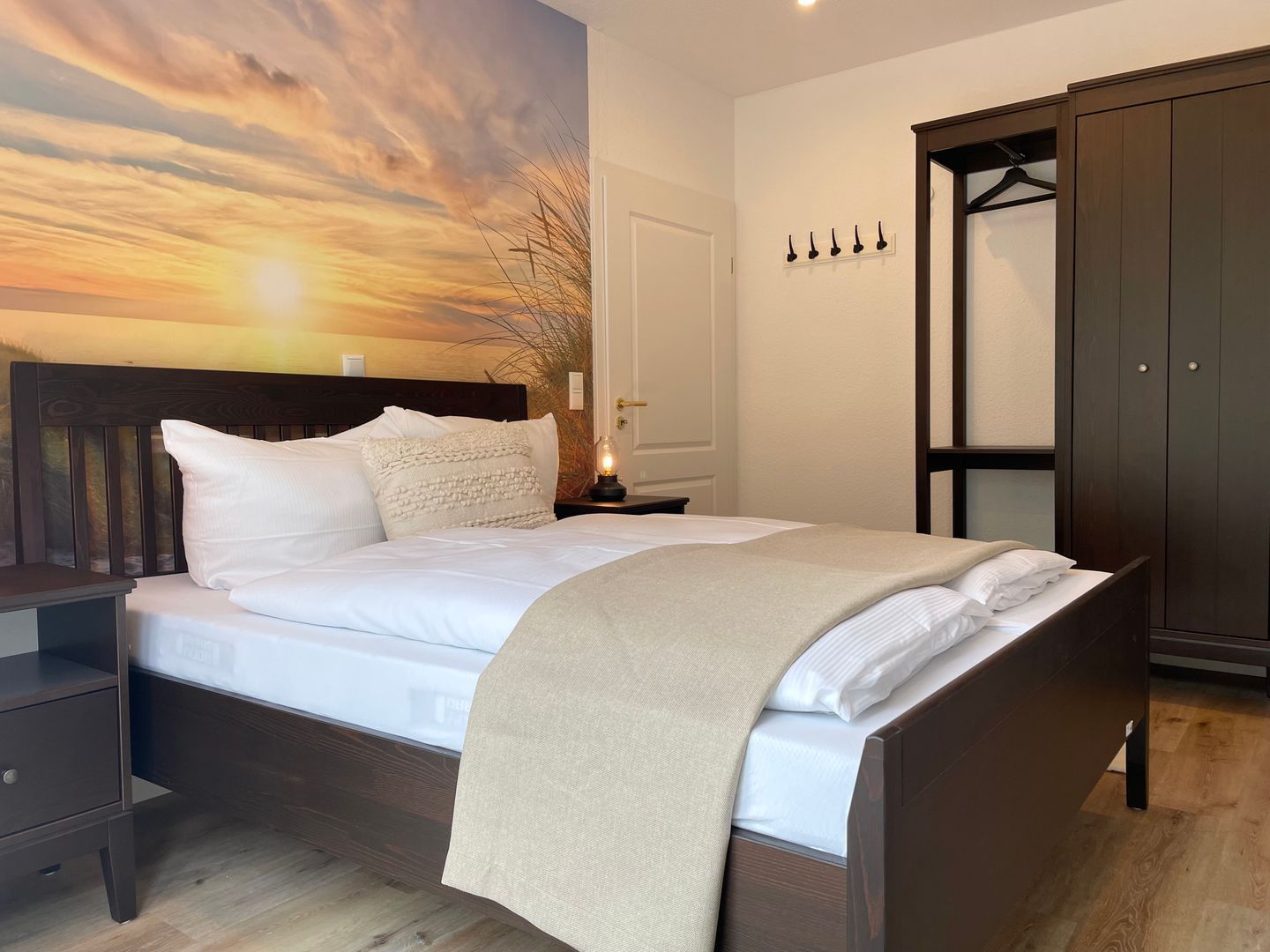 A bedroom with a bed, a nightstand, a wardrobe, and a wall with a sunset view.