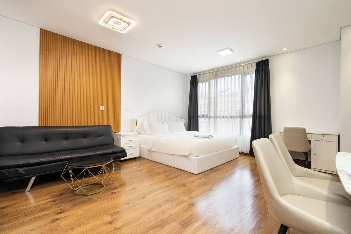 Ein modernes Hotelzimmer mit einer Wand in Holzoptik, einer schwarzen Couch, einem weißen Bett, einem Esstisch mit Stühlen und einem kleinen Beistelltisch.