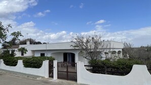 A modern white house with a gated entrance, surrounded by greenery and a clear sky.