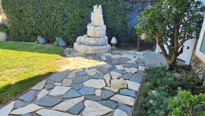 Un chemin de pierres menant à une fontaine en pierre, le tout étant entouré d’une haie et de diverses plantes.