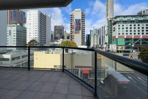 Paisaje urbano con edificios modernos, cielo despejado y vista desde un balcón con barandilla de vidrio.