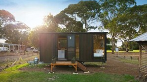 A modern, compact cabin with a large window, a small porch, and a view of a fenced area with trees.