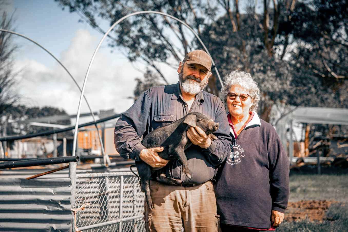 Two individuals holding a piglet in an outdoor setting with metal fencing and trees in the background.