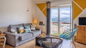 A modern living room with a grey sofa, a coffee table, and a blue chair. A TV is mounted on the wall, and there's a view of the city through the balcony doors. - Casa Sémaphore (L'Île-Rousse)