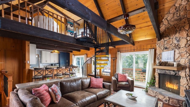 A spacious living area with a leather sectional sofa, a stone fireplace, and a staircase leading to an upper level. - Emerald Lake View Loft (Lake Arrowhead)
