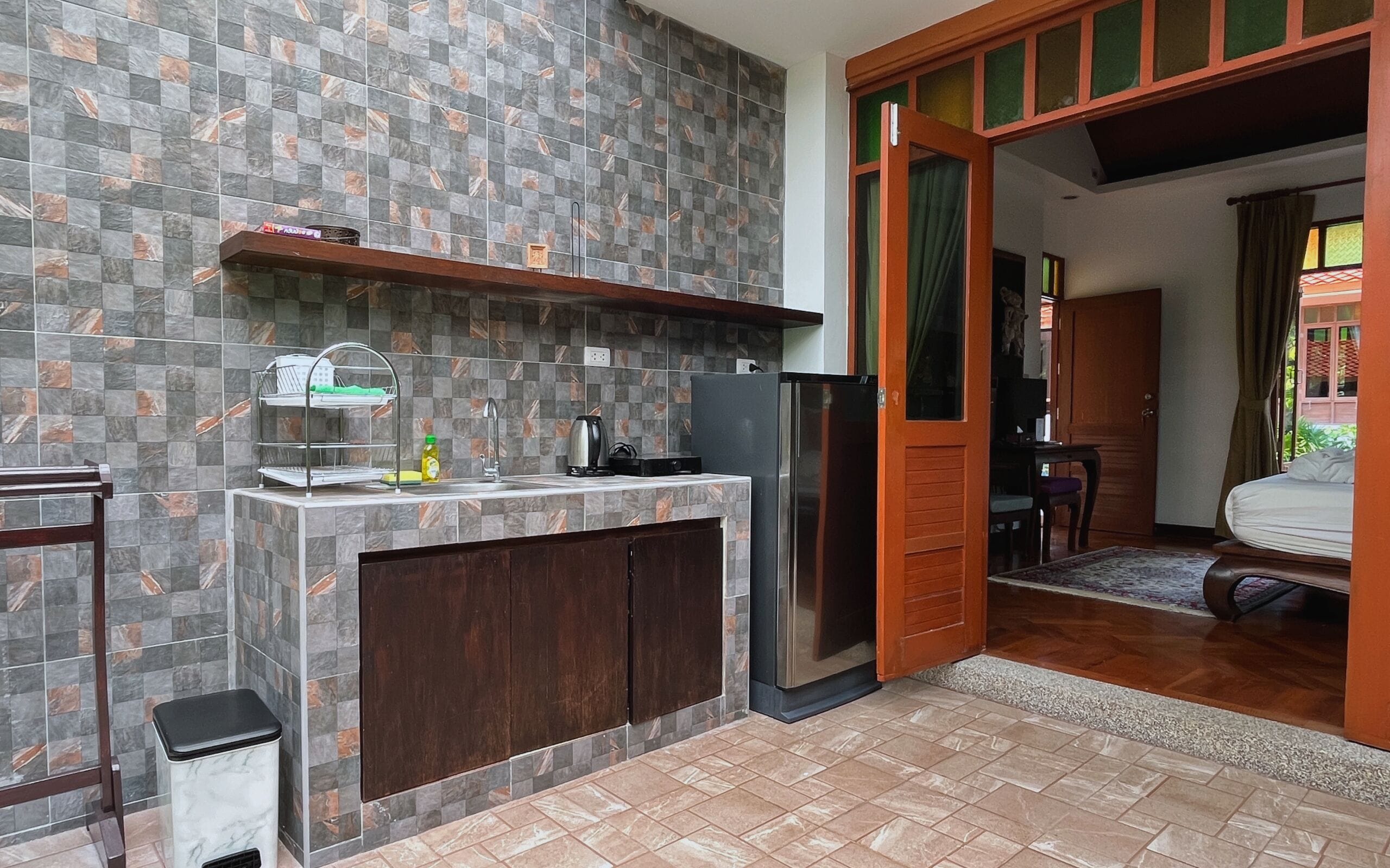 Une cuisine compacte équipée d’un réfrigérateur encastré, d’un évier et d’un petit plan de travail. Une porte en bois donne accès à une chambre à coucher qui contient un lit et une fenêtre.