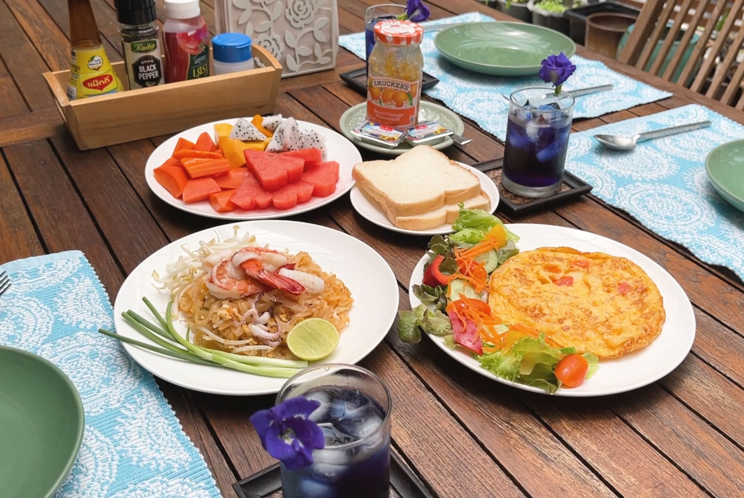 A wooden table with various dishes including noodles, omelette, and salad, along with condiments and drinks.