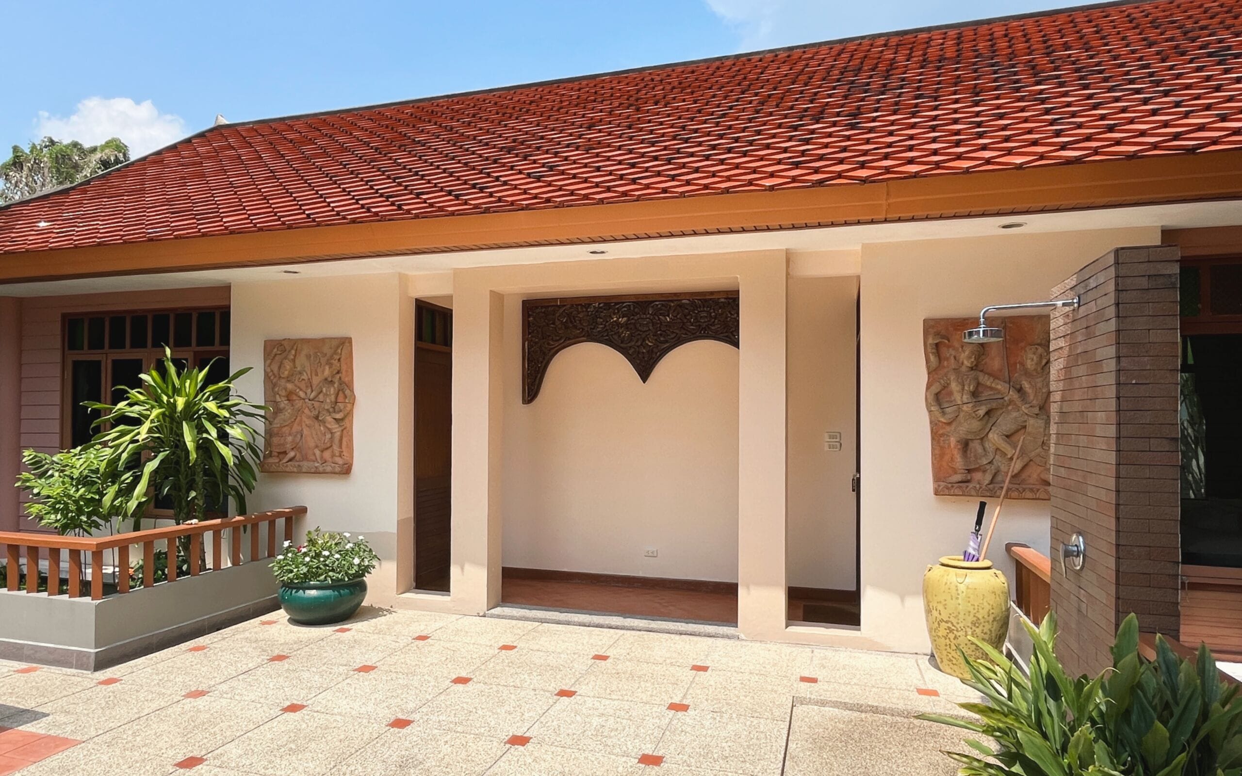 Un edificio con techo de tejas rojas, un vestíbulo cubierto con un arco decorativo y un patio con macetas y plantas.