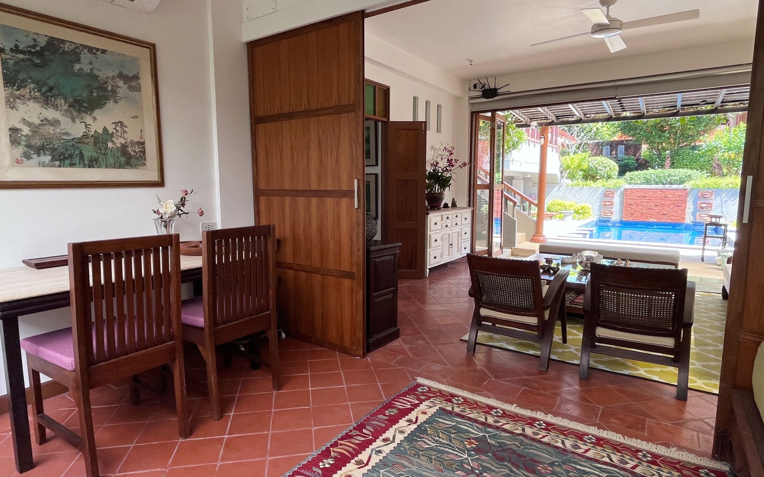 A dining area with wooden chairs and a table, a wooden partition, a cabinet with decorative items, and a view of an outdoor swimming pool.