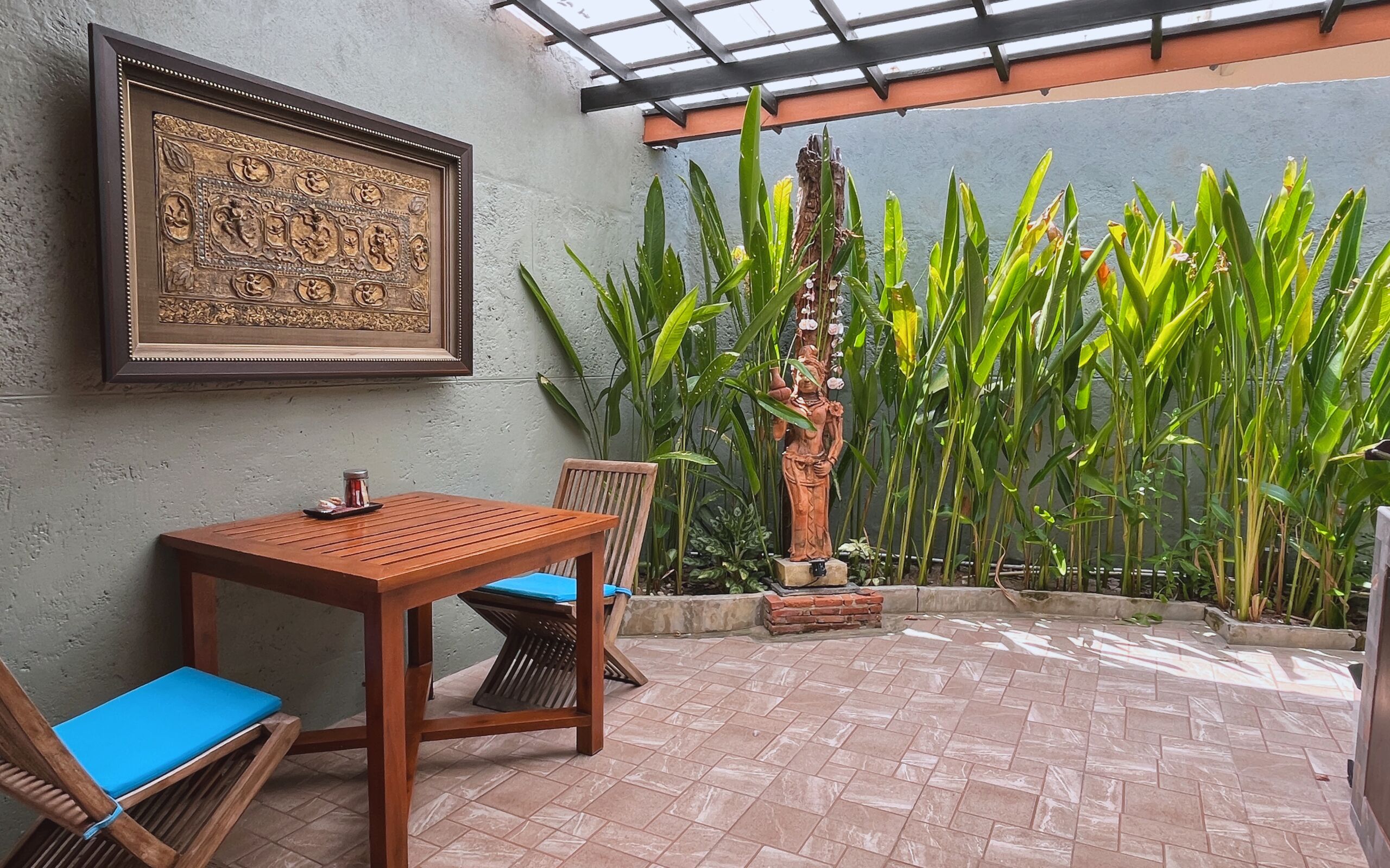 Eine Terrasse mit einem Holztisch und -stühlen, einem gerahmten Kunstwerk und hohen, grünen Pflanzen.