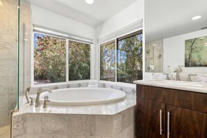 Une salle de bain moderne dotée d’une grande baignoire sur pieds, d’un meuble-lavabo double avec un plan de toilette en marbre et de grandes fenêtres offrant une vue sur les arbres.