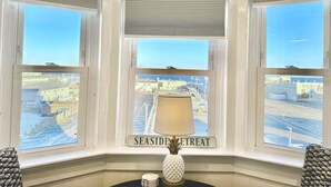 Quarto com três janelas amplas, uma mesa com abajur e uma placa com a inscrição 'SEASIDE RETREAT'.