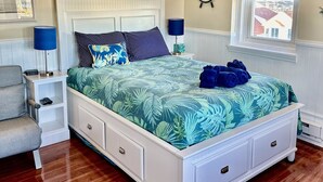 Quarto com cama, mesas de cabeceira, ventoinha no teto, janela com persianas e um leme fixado na parede.