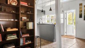 A modern kitchen with a central island, a wooden bookshelf filled with books, and a large window providing natural light. - 2 bedroom nice home in Karrebæksminde (Karrebæksminde)