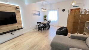En moderne stue med et fladskærms-tv monteret på en trævæg, en grå sofa, et spiseområde med bord og stole, samt en dekorativ plante.