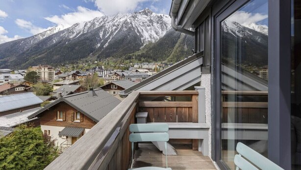Un balcón con vista a una cadena montañosa, bancos de madera y un edificio moderno con amplias ventanas.