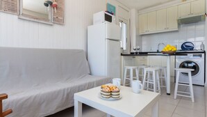 A compact living space with a sofa, a small table with a cake, a washing machine, and a kitchen area with stools and cabinets.