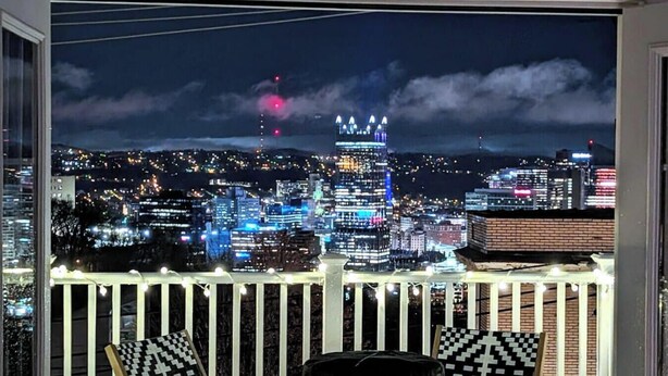 A cityscape at night with illuminated buildings and a balcony with string lights. - ★ HostWise Stays ★ Skyline City Views & Rooftop Bar at Mt. Washington (Pittsburgh)