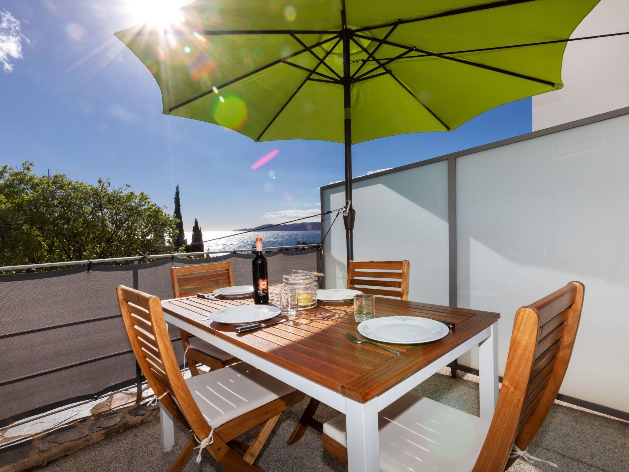 Een eettafel met stoelen, een fles wijn en glazen op een terras met uitzicht op zee.
