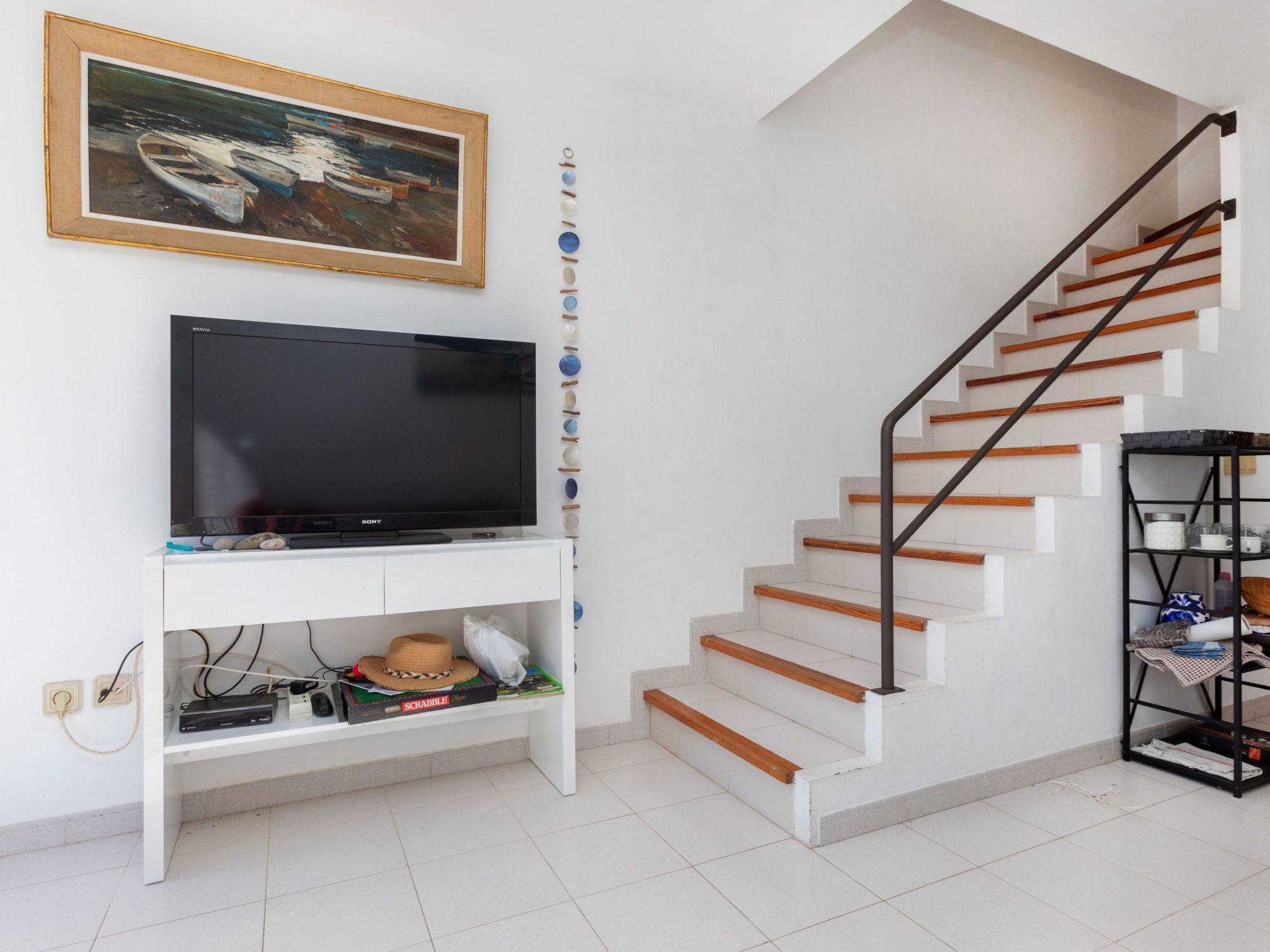 A staircase with wooden steps and a black handrail, a white entertainment unit with a flat-screen TV, and a shelf with various items including a hat and a board game.